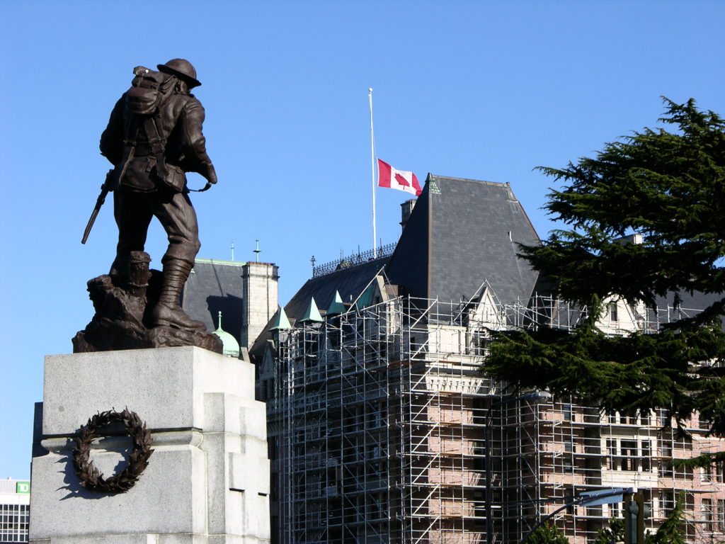 The Victoria War Memorial at the corner of Government and Belleville Streets. Image taken from behind the memorial looking upwards towards the soldier. In the background a Canadian flag flies at half-mast on the Empress Hotel which is covered with scaffolding.