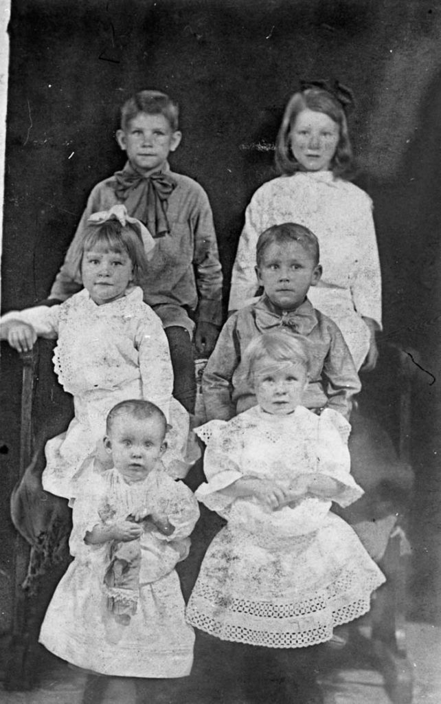 Six children of the Berget family. Four posed upon a large chair. The two boys wear large bows at their necks. The two older girls with ribbons in the hair. The youngest girls in the foreground are standing, the youngest holding a doll.