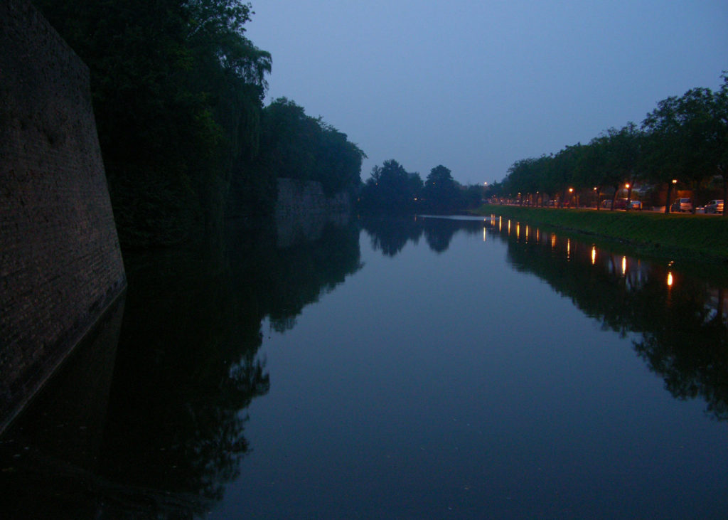 The canal outside the Menin Gate Memorial. It is after sunset. The sky is indigo blue-black and the earth's feature darker. Lights reflect off the canal in a row towards a vanishing point.