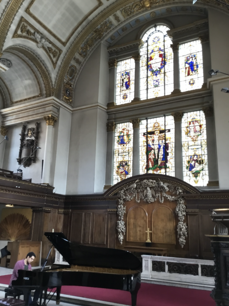 Inside St. James's Church a pianist plays at a Grand piano as light streams through the stained glass. The pianist plays in the altar area. It is a large area and one hopes that you can imagine the notes rising and falling within this space.