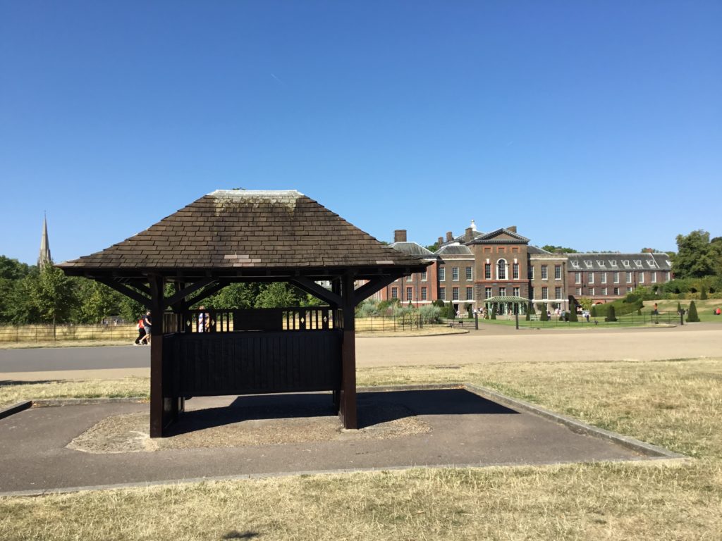 A wood shelter sits in an open space at Kengsington Gardens, London. Kensignton Palace in the background.