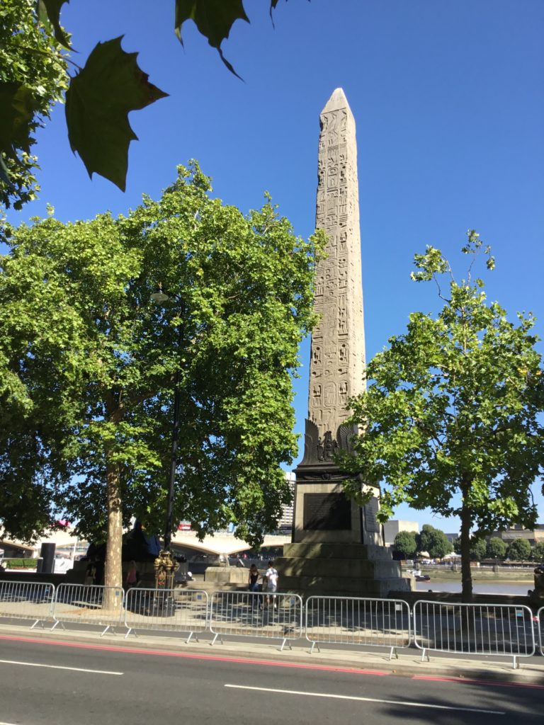 Cleopatra's Needle. The Thames in the background. A guard rail separates traffic along a middle median.