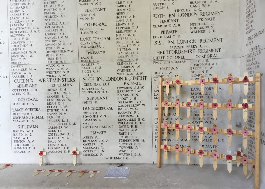 Panels of names on the Menin Gate War Memorial. Poppy crosses lay against the panels and at its base. In the corner a grid of poppy crosses 4 x 7 in number. One has detached itself. A note has been purposely left behind.