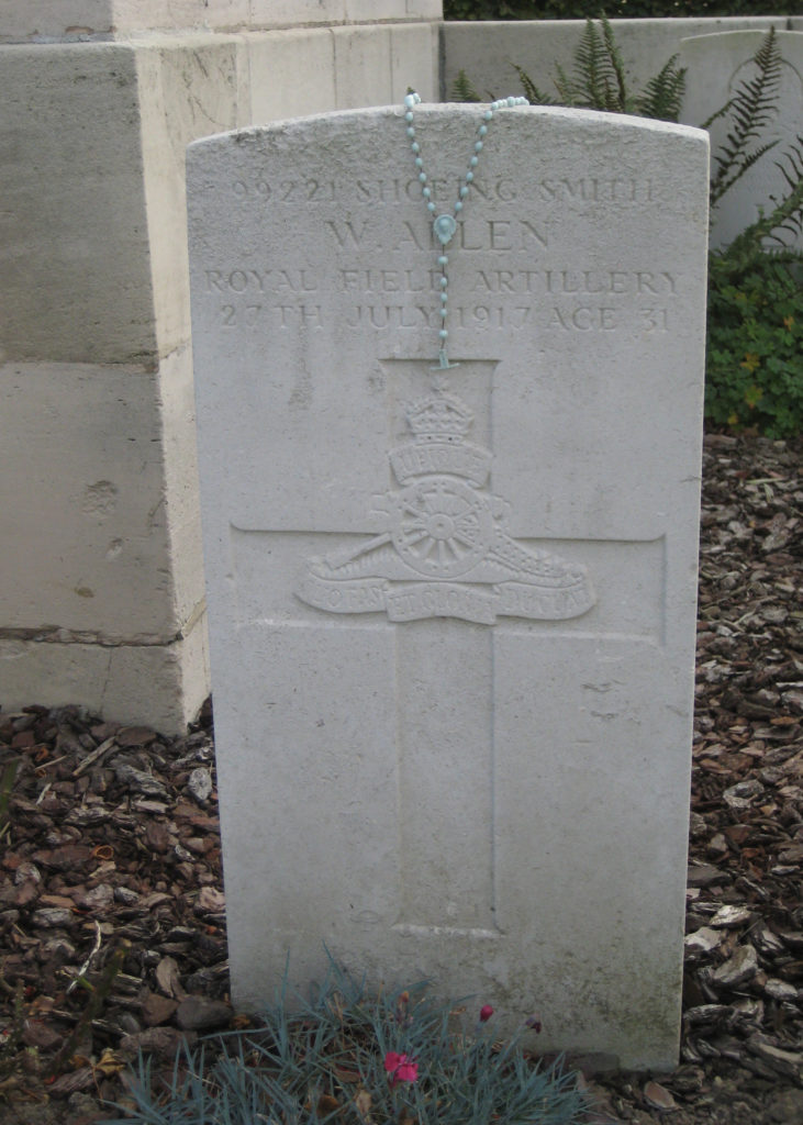 A light blue-green rosary is draped over the top of a Commonwealth War Graves Commission marker. W. Allen's name can be see with details above the insignia of the Royal Field Artillery emblazoned on a christian cross.