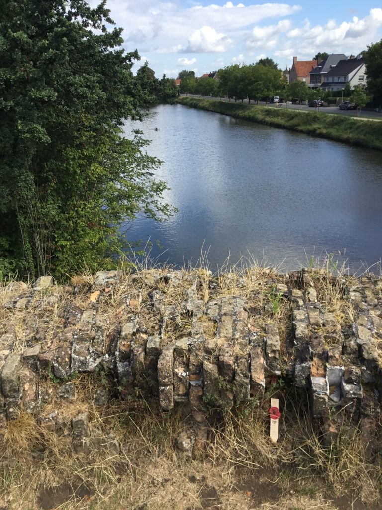 A non-denominational remembrance, made of wood and decorated with a red poppy rests against a low wall on the ramparts near Menin Gate. The canal in the background.