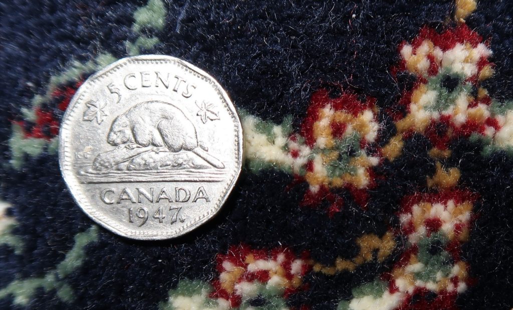 A 1947 Canadian nickel set against a carpeted background. The coin is multi-sided and shows a beaver atop its dam.