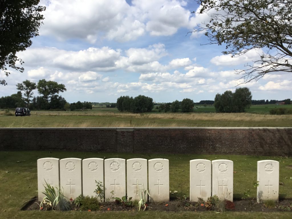 Nine Commonwealth War Graves Commission headstones at the back of Woods Cemetery, near to the wall. A van is in the field behind the wall. We are looking towards Ypres to a blue sky filled with puffy white clouds.