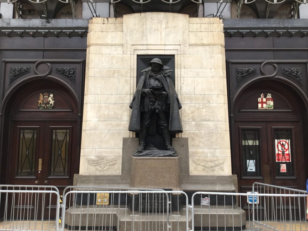 The Great Western Railway War Memorial at Paddington Station.  The soldier statue reading the letter framed by a white stone wall behind. He stands upon a plinth. There is a modern series of gates set up in front of the statue. Entry doors to either side of the memorial have the Royal Coats of Arms and another possibly relating to the City of London above the entries.