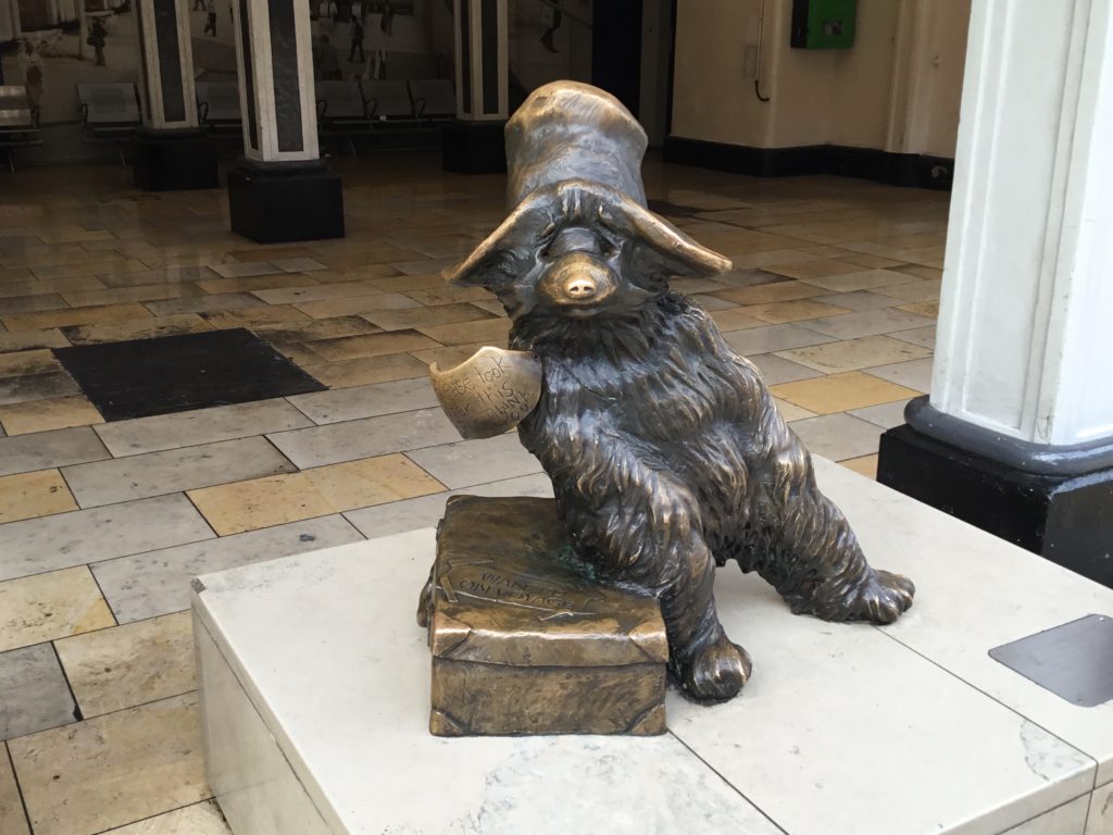 Bronze statue of Paddington Bear. Unlike the cartoon he does not wear a jacket or boots. He wears a large address label and large floppy hat. He sits on a suitcase.