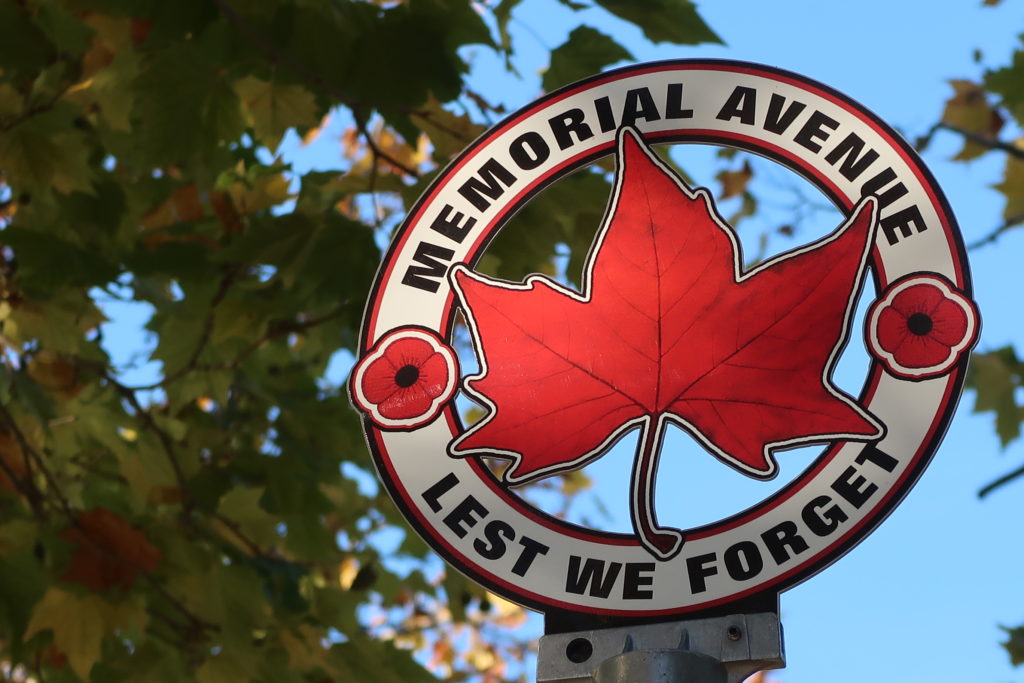 A circular sign atop a street sign-post. The colourful red, white and black sign features a red maple leaf and two red poppies. The sign reads "Memorial Avenue Lest We Forget".