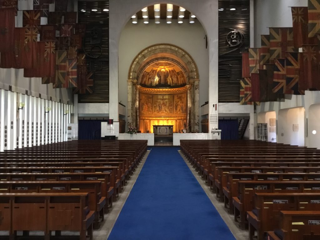 The Guards Chapel, London, England. Looking down the central aisle fitted with a deep blue carpet. Horizontal pews to wither side. Above the pews to the left and right, the Guard's colours hag from the wall. In the background the golden area of the chapel with altar.