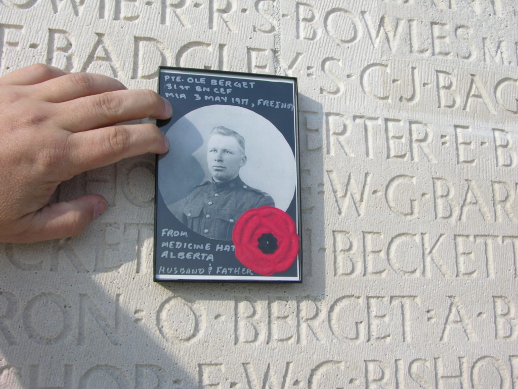 The writer's hand holds a framed portrait image of Private Ole Berget against the lettering of his name on the Vimy Memorial. There is a red poppy within the frame.
