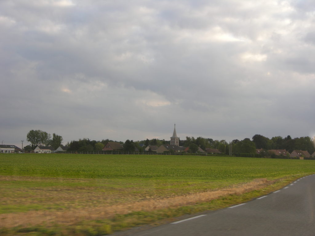 Looking towards the French village of Fresnoy. A green farmer's field in the foreground. On the horizon a church steeple rises above the town. It is a cloudy day.