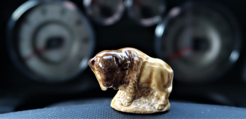 A china buffalo made by Wade stands upon a steering wheel with dashboard instruments in the background.